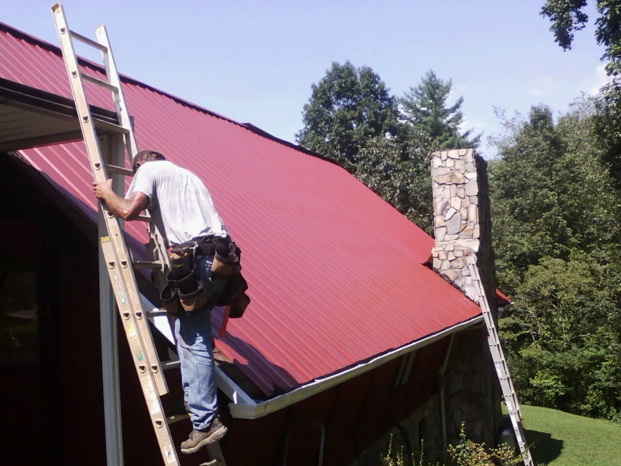 Roofer on a ladder beside a red metal roof and stone chimney — Soffit & Fascia Repair by Roofing Brothers in North Sioux City
